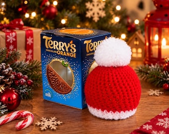 May include: A blue cardboard box of Terry's Chocolate Orange featuring a graphic of a chocolate segment, paired with a red crocheted hat cover topped with a white pom-pom and a white ribbed brim, designed to fit over the chocolate box.