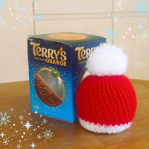 May include: A box of Terry's Chocolate Orange next to a small, handmade red and white Santa hat with a white pom-pom. The box is blue with the product name and a picture of the chocolate inside. The background is a light wood surface.