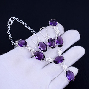 Könnte beinhalten: Ein silbernes Halsketten- und Ohrring-Set mit violetten ovalen Edelsteinen. Die Halskette hat eine Kette mit einem Verschluss und die Ohrringe sind Ohrhänger.