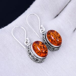 Op de afbeelding: Een paar zilveren oorhangers met ovale amberstenen. De stenen zijn diep oranje van kleur met een lichte bruine tint. De oorhangers hebben een delicaat, ingewikkeld ontwerp.