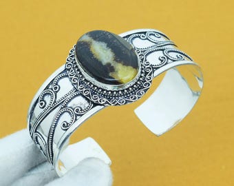 May include: Silver cuff bracelet featuring an oval, dark brown and yellow gemstone. The bracelet has an open design with intricate scrollwork and a polished finish. The gemstone is set in a detailed bezel.