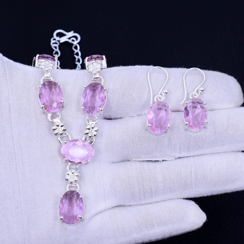 May include: A silver necklace and earrings set with pink oval-shaped gemstones. The necklace features a delicate chain with floral accents and a dangling pendant. The earrings are drop style with a single gemstone.