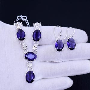 May include: A silver necklace and earrings set with oval-shaped purple gemstones. The necklace has a delicate chain with floral accents and three gemstones. The earrings are simple studs with a single gemstone.