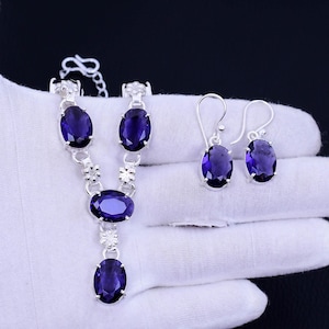 May include: A silver necklace and earrings set with oval-shaped purple gemstones. The necklace has a delicate chain with floral accents and three gemstones. The earrings are simple studs with a single gemstone.
