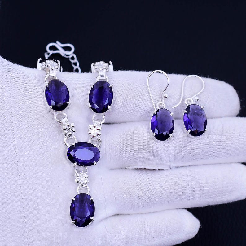 May include: A silver necklace and earrings set with oval-shaped purple gemstones. The necklace has a delicate chain with floral accents and three gemstones. The earrings are simple studs with a single gemstone.