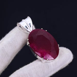 Puede incluir: Un colgante de plata con una gran piedra preciosa de rubí de forma ovalada. El rubí es de color rojo intenso y tiene un corte facetado.