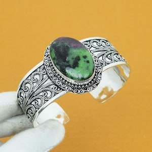 以下が含まれることがあります： 緑とマルーン色の楕円形の宝石をあしらったシルバーのカフブレスレット。ブレスレットは、バンドと宝石の周りに、精巧なスクロールワークのデザインが施されています。宝石の長さは約2.5cmです。