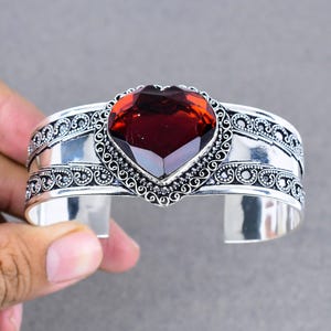 May include: A silver cuff bracelet with an ornate design and a large, red heart-shaped gemstone.