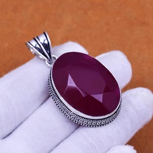 May include: A large, oval-shaped pendant with a faceted, deep red gemstone. The gemstone is set in a silver-colored metal frame with an ornate design. The pendant has a bail for attaching to a chain. The background is a solid, warm brown color.