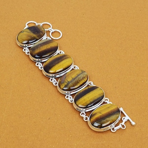 May include: A silver bracelet featuring six oval tiger's eye stones. The stones are a mix of yellow and brown with a striped pattern. The bracelet has a clasp closure and is set on a brown background.
