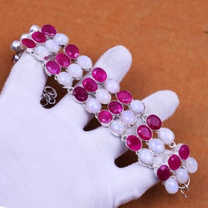 Op de afbeelding: Een zilveren armband met afwisselende ovale robijn- en maansteen edelstenen. De armband heeft drie rijen edelstenen en een veilige sluiting. De edelstenen zijn gezet in een zilveren metalen zetting.