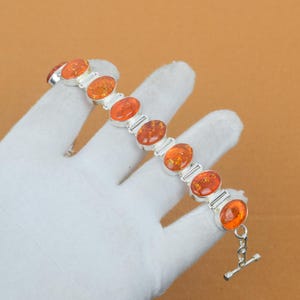 Peut inclure: Un bracelet en argent orné de pierres d'ambre ovales. Les pierres d'ambre sont de couleur orange chaud et serties dans du métal argenté. Le bracelet est doté d'un fermoir à bascule.