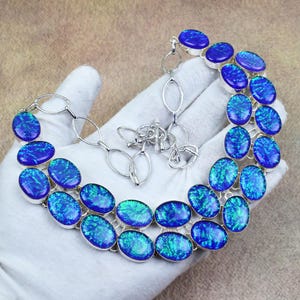 Op de afbeelding: Een ketting met twee rijen ovale blauwe en groene iriserende stenen in een zilverkleurige zetting. Het sieraad is voorzien van een zilverkleurige ketting met ovale schakels en een staafsluiting, met een levendig kleurenspel in de stenen.