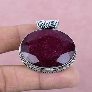 Può includere: Un ciondolo d'argento con una grande pietra di rubino rosso scuro sfaccettata. Il ciondolo ha una balla d'argento decorata.