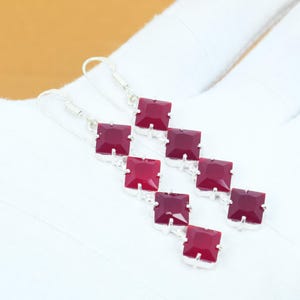 May include: A pair of dangling earrings featuring square, deep red gemstones set in silver-colored metal. The earrings have a linear design with multiple gemstones cascading down. The gemstones are a vibrant, rich red color.