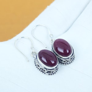Puede incluir: Un par de pendientes colgantes plateados. Cada pendiente presenta una gema ovalada de color rojo oscuro engastada en un marco decorativo de estilo filigrana. Los pendientes se muestran sobre un fondo blanco.