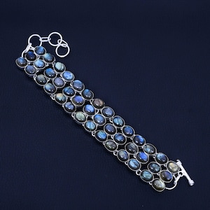 Könnte beinhalten: Ein silbernes Armband mit drei Reihen ovaler Labradoritsteine. Die Steine zeigen blaue, grüne und graue Farbtöne. Das Armband hat einen Knebelverschluss und eine Kette zur Größenanpassung.