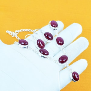 May include: A silver necklace and earring set featuring oval, deep red gemstones. The gemstones are set in a silver metal setting and connected by silver chains. The jewelry is displayed on a white surface against a yellow background.