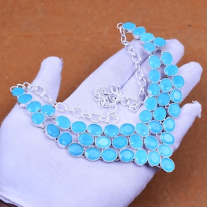May include: A multi-layered necklace with oval, light blue gemstones set in silver-coloured metal. The necklace has a chain fastening and is displayed on a white surface. The gemstones are arranged in a cascading pattern.