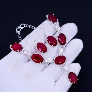 Könnte beinhalten: Eine silberne Halskette und Ohrringe mit roten ovalen Edelsteinen. Die Halskette hat einen Anhänger mit einem einzigen roten Edelstein.