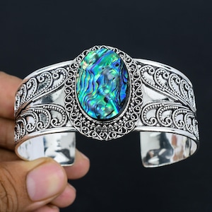 Puede incluir: Pulsera de brazalete de plata con una incrustación ovalada de concha de abulón. La pulsera tiene un diseño decorativo con espirales y volutas.