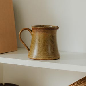 Może przedstawiać: Ręcznie wykonany ceramiczny dzbanek z uchwytem i dzióbkiem. Dzbanek ma nakrapianą, ziemistobrązową glazurę z ciemniejszymi brązowymi akcentami przy krawędzi. Stoi na białej półce.