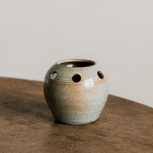 Könnte beinhalten: Eine kleine, runde Keramikvase mit hellblau-brauner, gesprenkelter Glasur. Die Vase hat eine Reihe von gleichmäßig verteilten Löchern um die Oberseite und steht auf einer Holzoberfläche. Die Vase ist ein dekorativer Gegenstand.