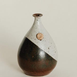 Może przedstawiać: Ręcznie wykonany ceramiczny wazon o unikalnym designie. Wazon posiada ukośny blok kolorów z ciemnobrązowym i białym nakrapianym wykończeniem. Mały, okrągły, drewniany akcent jest przymocowany do białej części wazonu.