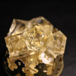 Puede incluir: Un conjunto de dados transparentes de color dorado con números visibles en el interior, que se encuentran en un soporte de metal dorado con un diseño de ancla.