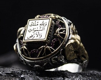May include: A silver ring with gold accents and a square centerpiece featuring Arabic script. The ring has an ornate design with small red stones and elephant head details. The ring is set against a dark background.