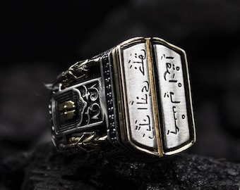 May include: A silver ring with gold accents and Arabic script. The ring has a rectangular face with a gold border and is set on a dark surface. The ring's design includes intricate patterns and a contrasting color scheme.