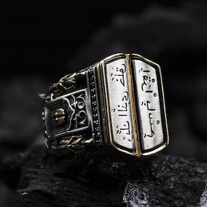 May include: A silver ring with gold detailing and Arabic script. The ring features a rectangular face with a gold border, set against a dark backdrop. The design incorporates detailed patterns and a contrasting colour scheme.