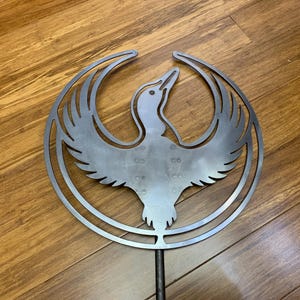 May include: A silver metal bird sculpture with outstretched wings, set within a circular frame. The bird is depicted in a stylized, minimalist design. The sculpture is mounted on a dark metal rod, suitable for garden or yard decor.