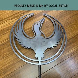 May include: A metal bird sculpture with outstretched wings, set within a circular frame. The sculpture is made of silver-colored metal and is mounted on a black rod. The text "PROUDLY MADE IN MN BY LOCAL ARTIST!" is at the top.