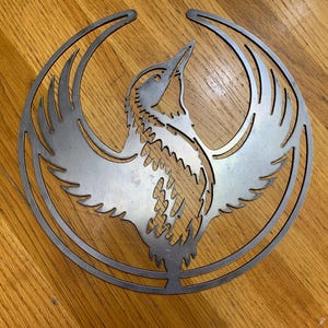Puede incluir: Una pieza de arte mural de metal que representa un pájaro estilizado con las alas extendidas, dentro de un marco circular. El diseño del pájaro es intrincado, con patrones de plumas detallados. La obra de arte es de metal plateado.