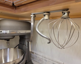 Op de afbeelding: Een zilveren standaard met twee haken om keukenmixeraccessoires aan op te hangen. Een zilveren garde en een zilveren deeghaak hangen aan de haken. De standaard is gemonteerd onder een houten kast.