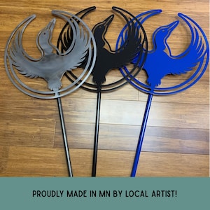 May include: Three metal garden stakes, each featuring a circular frame with a cutout silhouette of a loon in flight. The stakes are finished in metallic silver, matte black, and vibrant blue. The text PROUDLY MADE IN MN BY LOCAL ARTIST is visible.