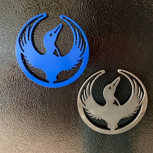 May include: Two circular emblems, one blue and one silver, each featuring a stylized bird within a ring. The bird has outstretched wings and a long beak. The emblems are set against a dark, textured background.