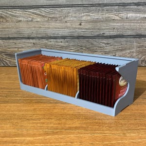 May include: A gray plastic tea bag organizer with three compartments. The compartments are filled with tea bags in different colors, including brown, orange, and red.