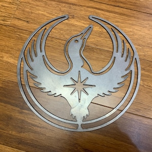 May include: A silver metal wall art piece featuring a stylized bird with outstretched wings and a star in the center. The bird is enclosed within a circular frame. The artwork is displayed on a wooden surface.