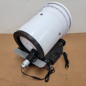 May include: A white plastic bucket mounted on a gray metal frame with a black gear ring attached to the rim. A small electric motor with a gear is connected to the ring, powered by a black power adapter with a cord and a standard two-prong plug.