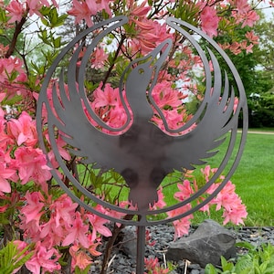 May include: A dark metal bird sculpture with outstretched wings, framed by a circular design. The sculpture is placed in a garden with vibrant pink flowers and green foliage in the background. The sculpture is mounted on a metal rod.