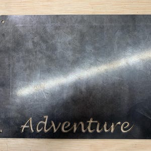 May include: A rectangular metal sign with the word "Adventure" cut out in a cursive font. The sign is dark gray with a textured surface and has four holes in the corners for mounting.