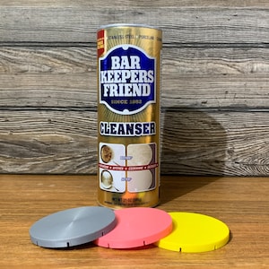 May include: A gold can of Bar Keepers Friend Cleanser with the text "Stainless Steel Porcelain Ceramic" and "Since 1882". The can is sitting on a wooden surface with three round, colorful coasters: gray, pink, and yellow.