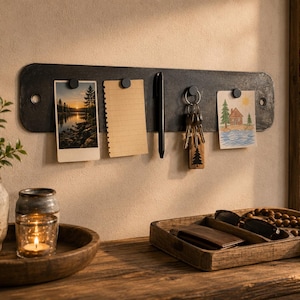 May include: A rectangular dark metal wall-mounted magnetic organiser strip featuring four small circular magnets. It holds a landscape photograph, a blank lined notepad, a black pen, a set of keys on a ring, and a small drawing of a house.