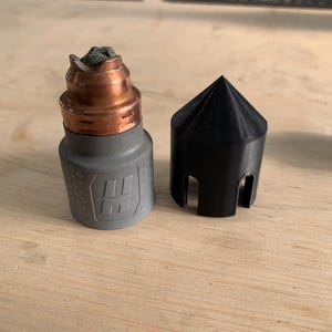 Puede incluir: Un dispositivo de vapeo gris y cobrizo con una tapa negra. El dispositivo tiene un cuerpo cilíndrico con un logotipo y una parte superior de cobre. La tapa negra tiene forma de cono con recortes. Los artículos están sobre una superficie de madera.