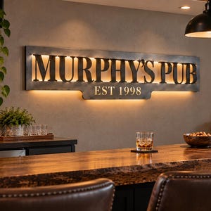May include: A rectangular metal wall sign featuring the cut-out text MURPHY'S PUB and EST 1998, illuminated from behind with a warm glow. The sign has a dark, weathered finish and is mounted horizontally on a neutral-toned wall above a wooden bar.