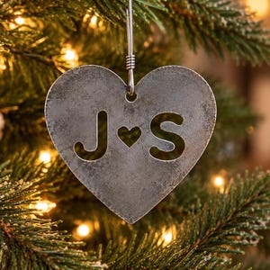 May include: A heart-shaped metal ornament with a weathered gray finish featuring the cut-out letters J and S separated by a small heart symbol. The ornament hangs from a silver wire loop against a background of green pine branches.