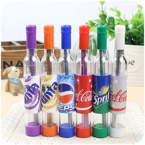May include: Six clear glass bottles with colorful lids and labels. The bottles contain pencils and have different brand logos, including Sprite, Coca-Cola, Pepsi, and Fanta.