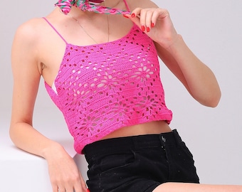 May include: A bright pink, crocheted crop top with a floral pattern. The top has thin straps and is paired with black shorts. A patterned scarf is around the neck. The model is sitting on a white surface.